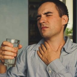 A parched throat and a glass of water in hand. Handsome guy in casual clothes is holding a glass of water and touching his throat while sitting on couch at home.
